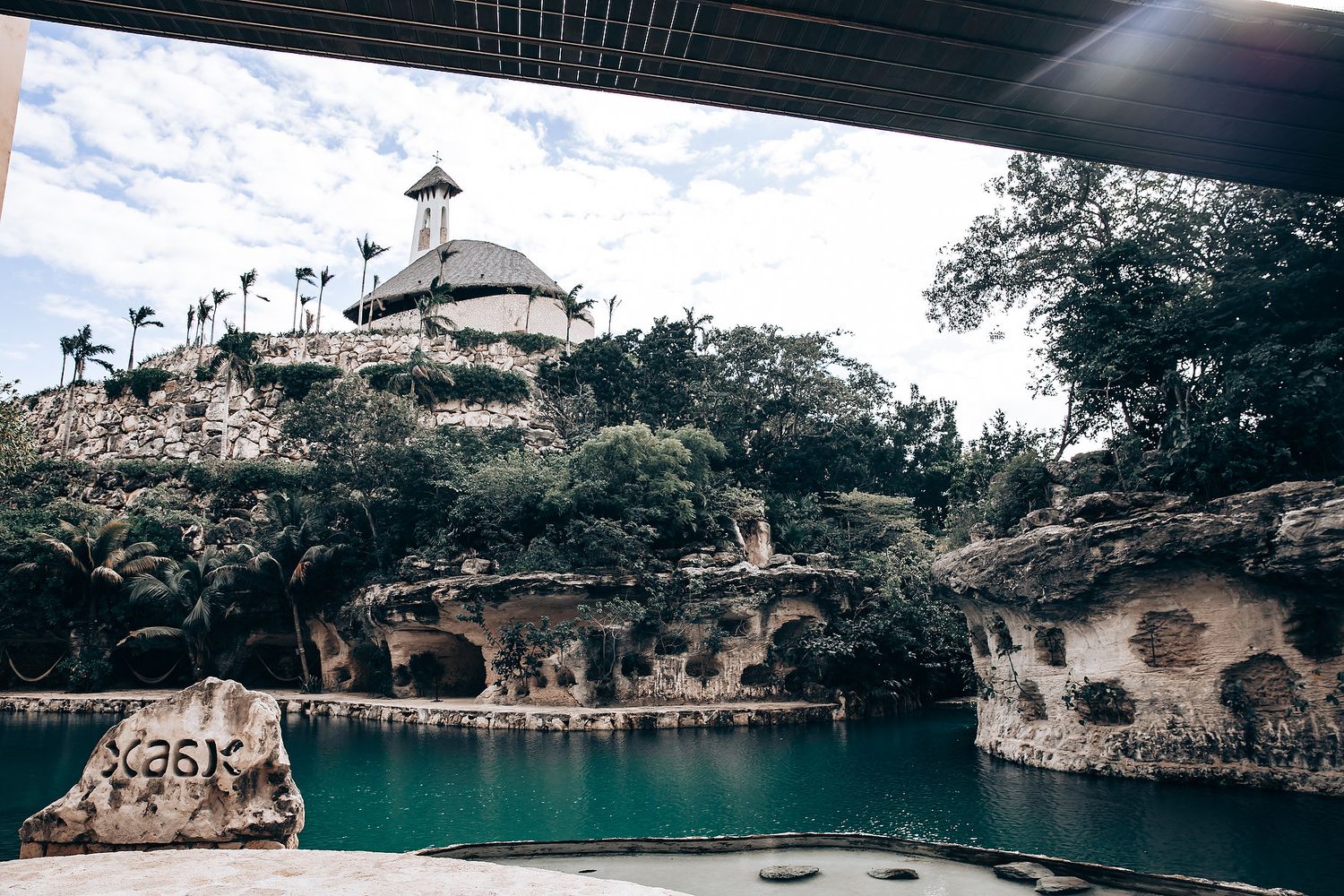Tropical resort pool with thatched roof gazebo and rock formations surrounded by palm trees.