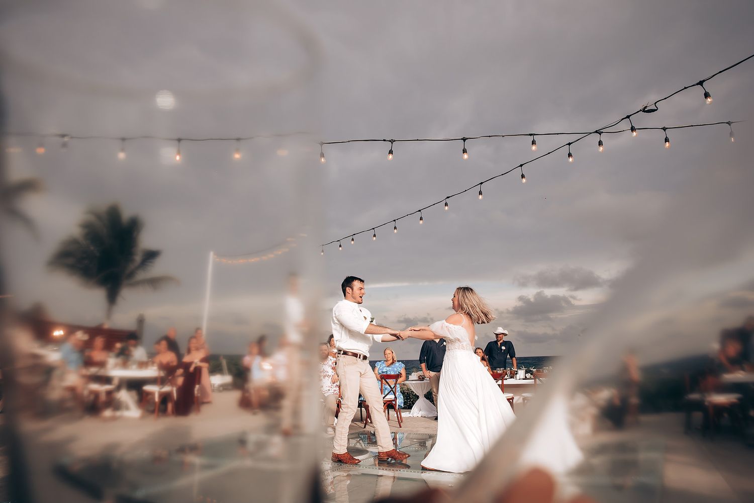 String lights illuminate an outdoor evening celebration with palm trees and a dreamy sunset backdrop.