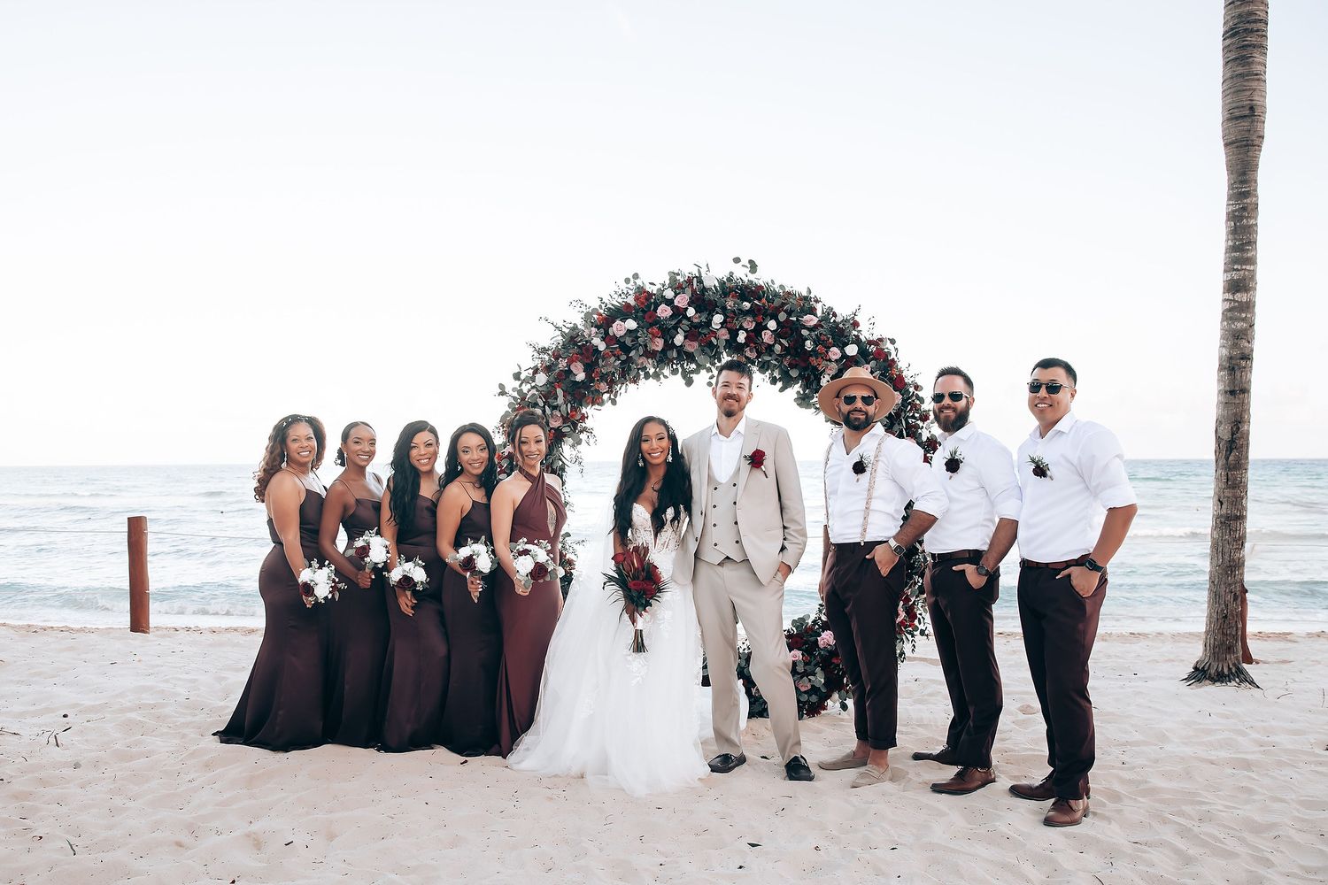 Beach wedding party photo with palm trees and ocean in the background.