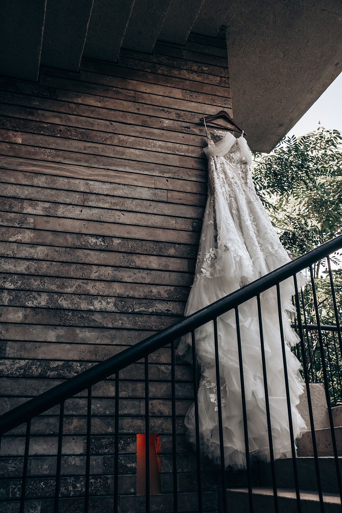 A white lace wedding dress hangs elegantly against rustic wooden siding with natural lighting filtering through.