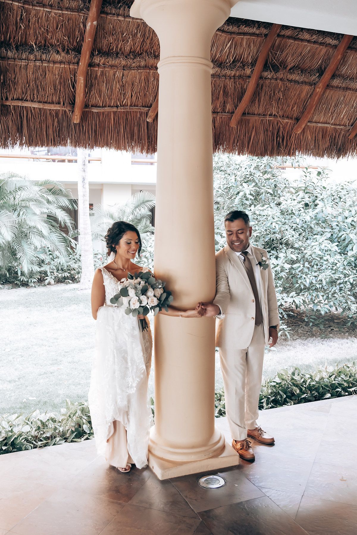 A couple poses under a thatched roof pavilion with tropical foliage in the background.