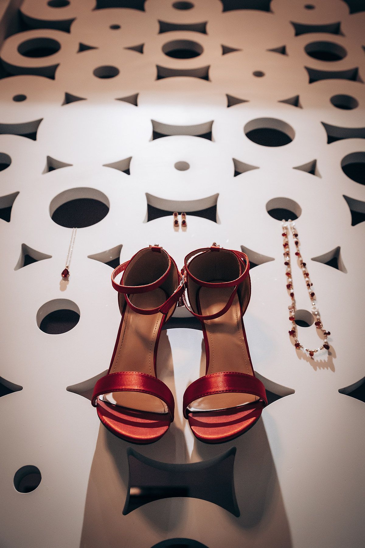 Red metallic sandals and jewelry displayed on a white geometric surface with circular patterns.