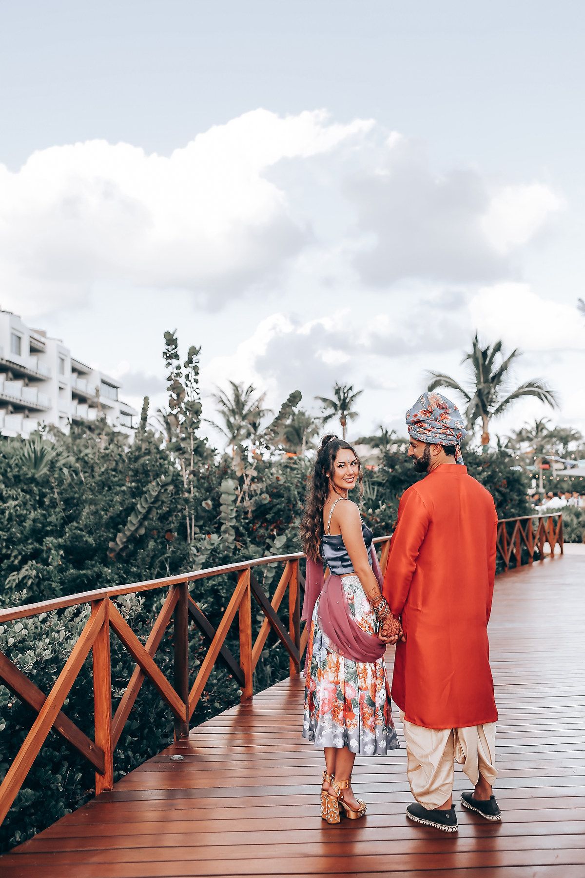 Couple strolling along a tropical wooden boardwalk with palm trees and buildings in the background.