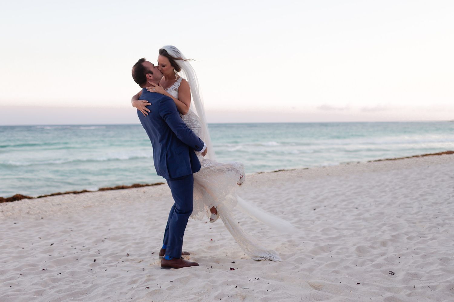 A romantic couple embraces on a sandy beach at sunset while waves roll in from the ocean.