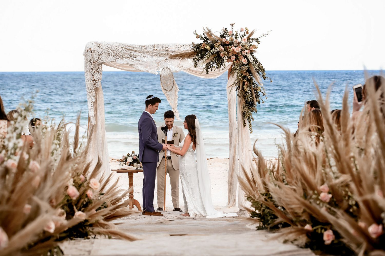 Beachfront wedding ceremony setup with dried palm fronds and ocean view.