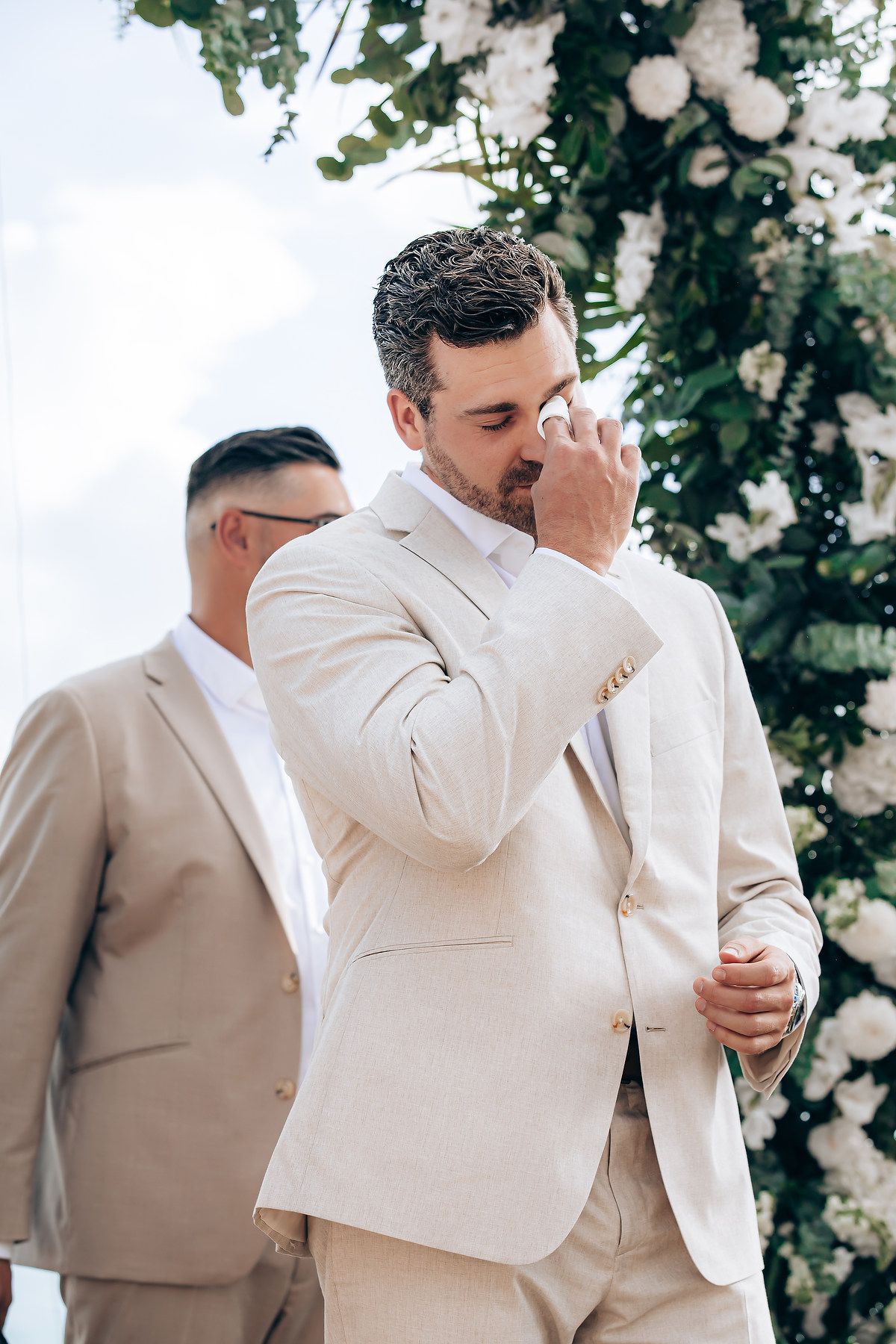 Groomsmen in light beige suits stand near a floral arch during a wedding ceremony.