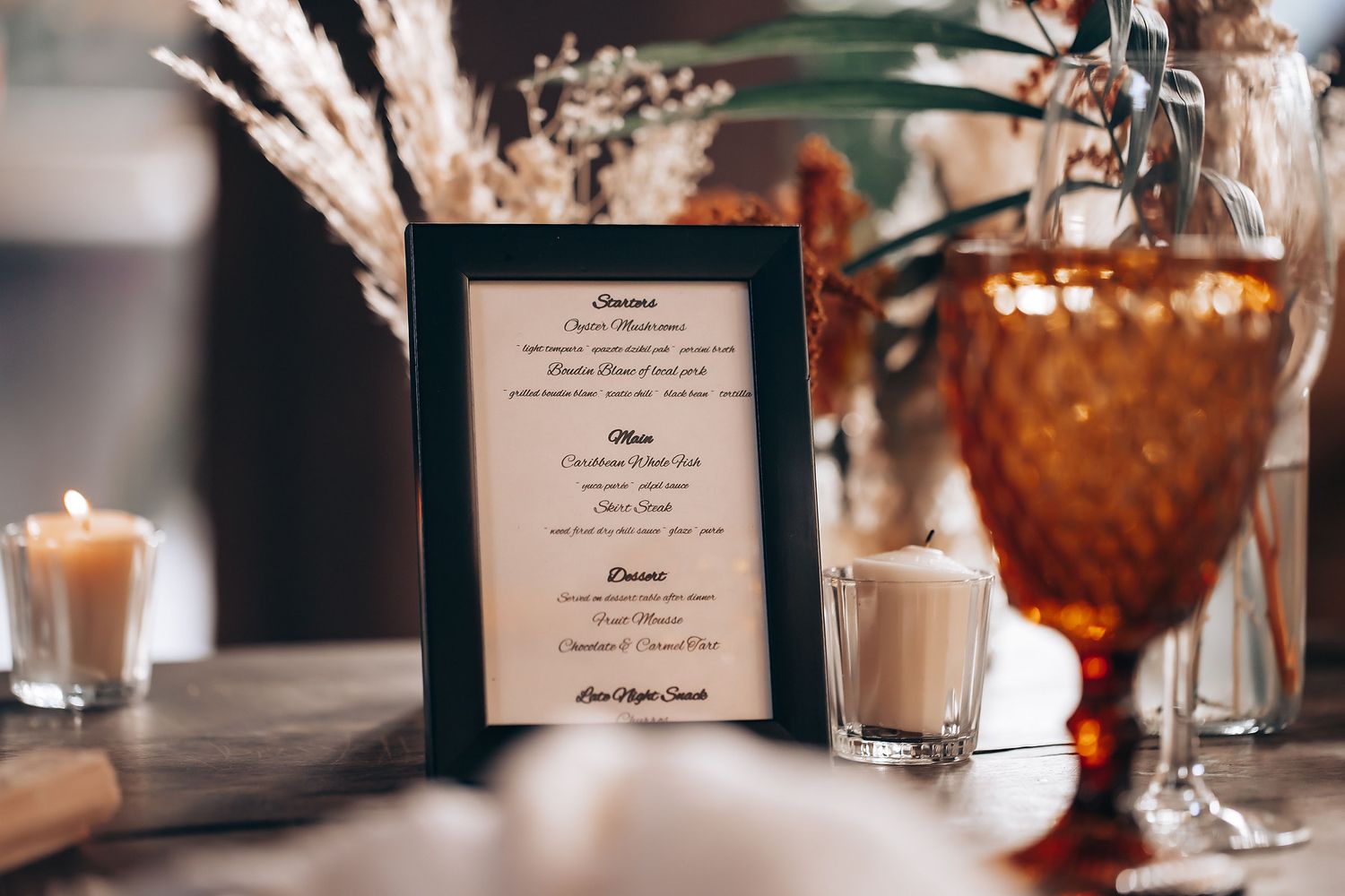 Elegant wedding reception table settings with amber glassware, candles, and menu cards in warm lighting.