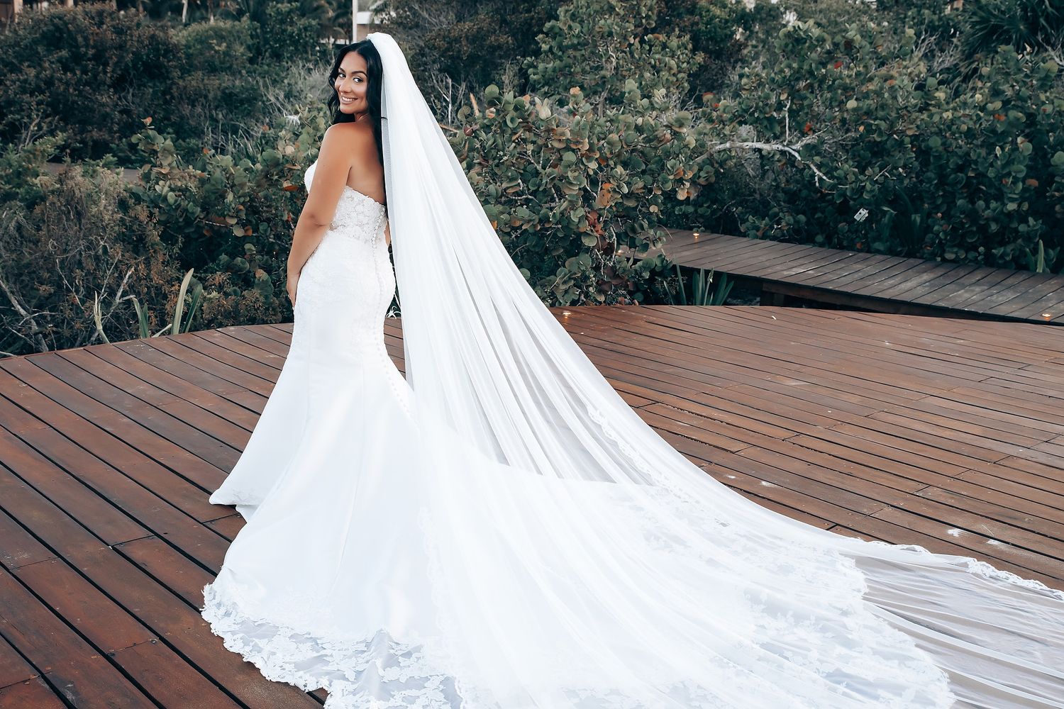 A flowing white wedding dress train trails across a wooden deck surrounded by lush greenery.