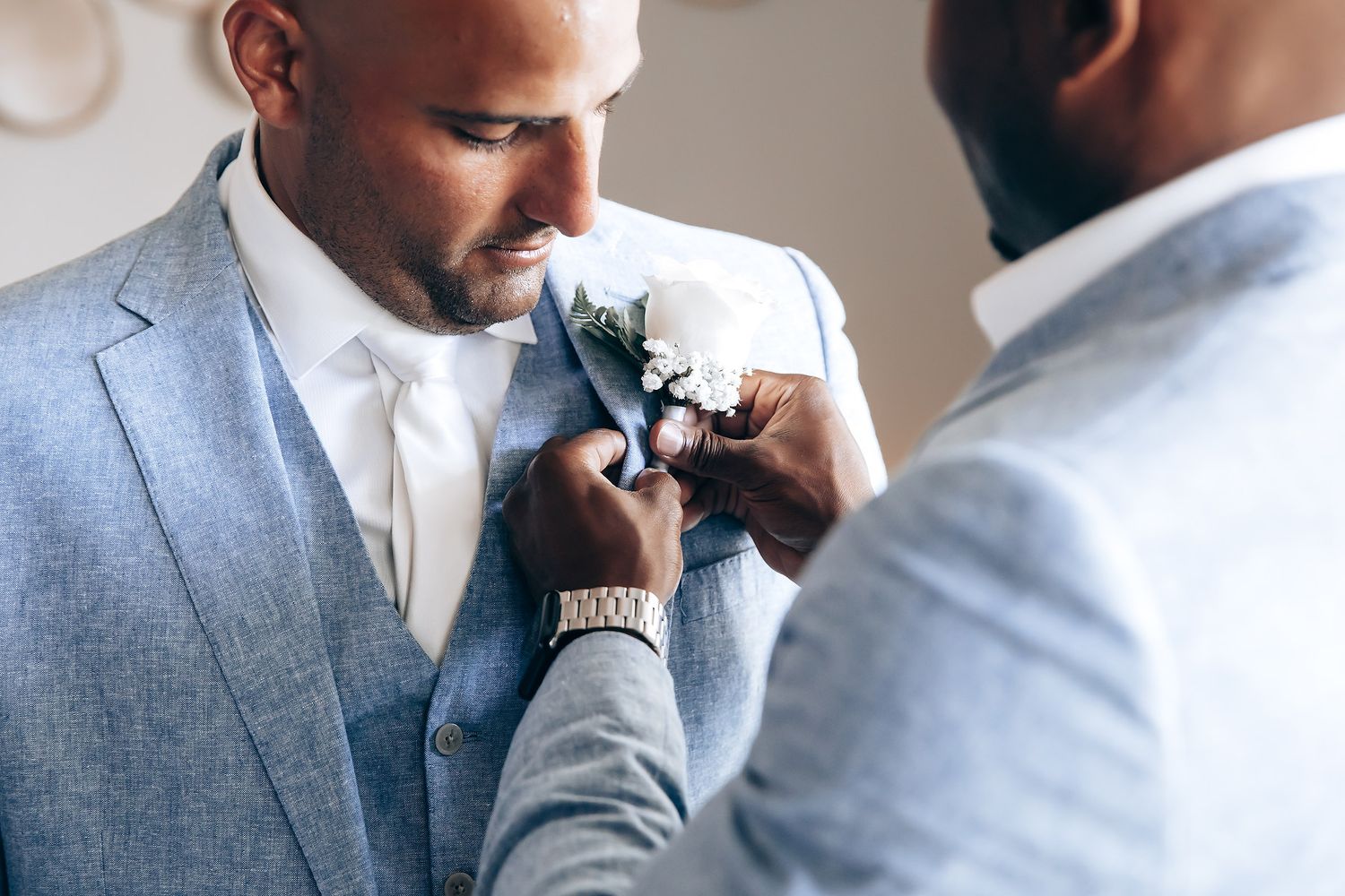 A groom adjusts his bowtie while getting ready in a light-filled room wearing a classic grey suit.