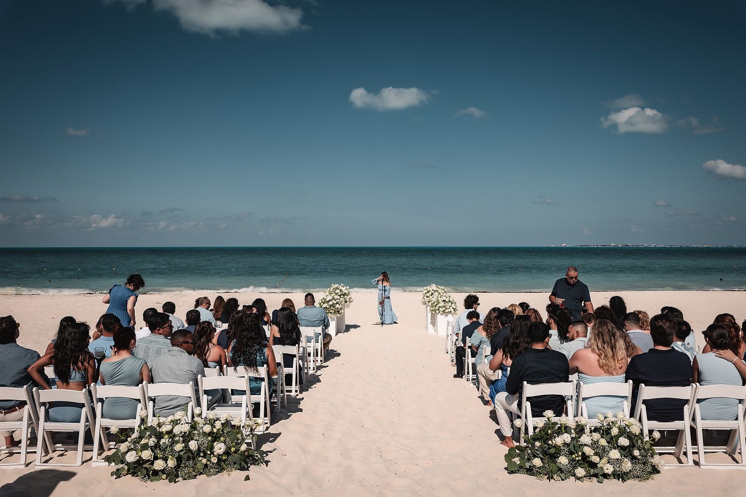 Beach wedding ceremony setup with white chairs and guests on a pristine sandy shoreline.