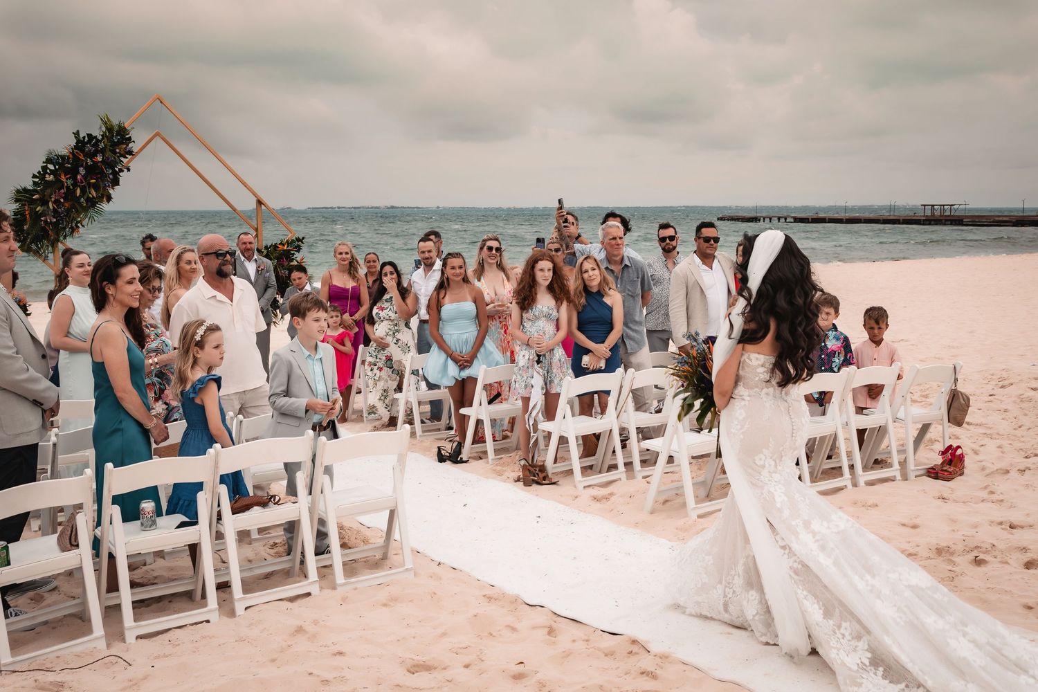 Wedding guests gather on a beach under cloudy skies for an oceanfront ceremony.