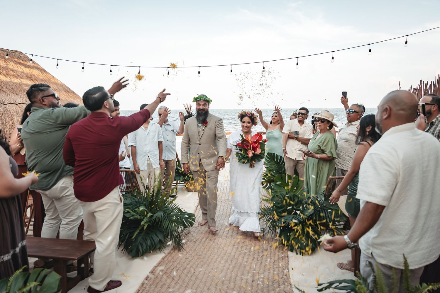 Joyful wedding couple walks down the aisle as guests celebrate with confetti at an outdoor beach ceremony.