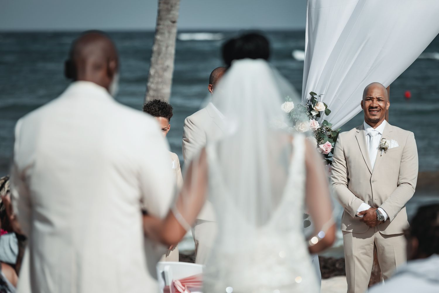 Groom smiling at bride during outdoor beach wedding ceremony with white draping and ocean backdrop.