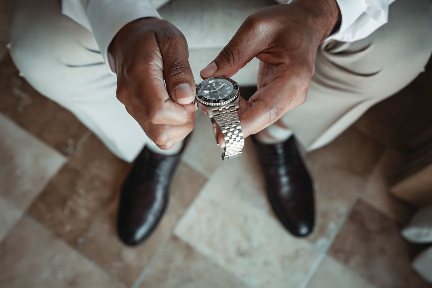 Groom holding a silver watch above brown dress shoes on marble floor, overhead view.