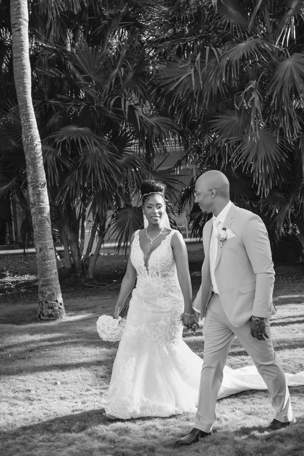 Black and white photo of a bride and groom walking hand in hand among tropical palm trees on their wedding day.