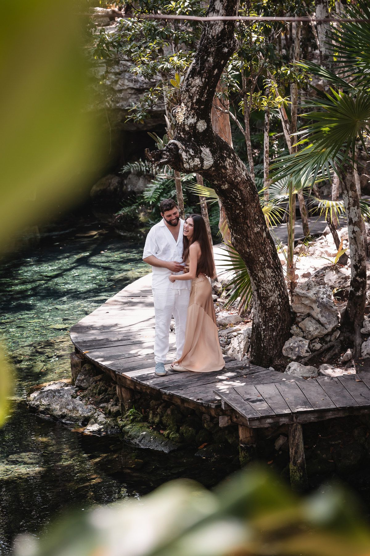 Romantic couple embracing on a wooden dock beside a crystal-clear cenote in a tropical jungle.