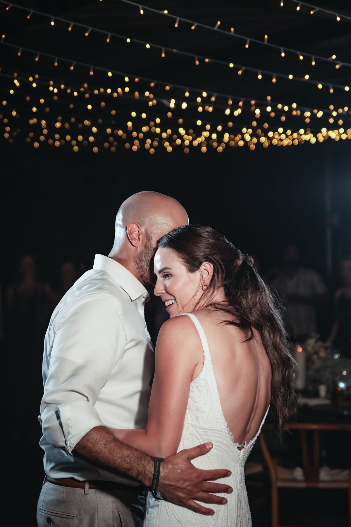 Couple sharing first dance at wedding reception under warm bokeh string lights at night.