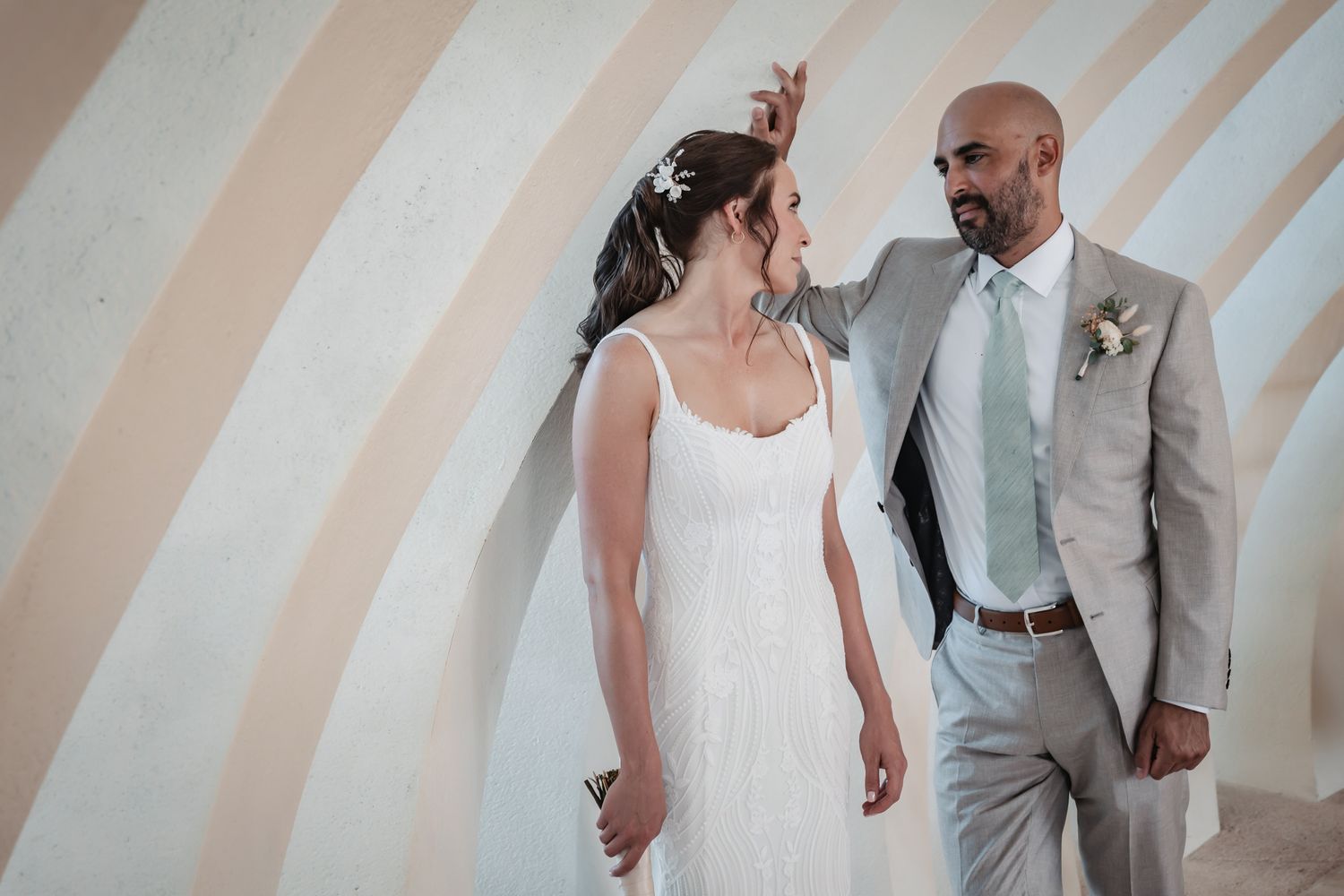 Bride and groom share a romantic moment against a striped wall, she in a white gown, he in a gray suit.