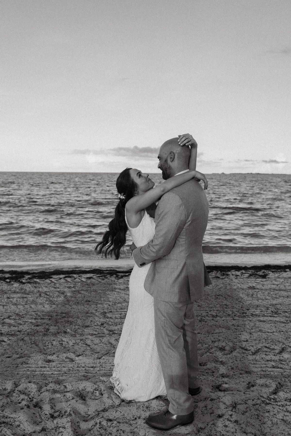Black and white photo of newlyweds embracing on a sandy beach with ocean waves in the background.