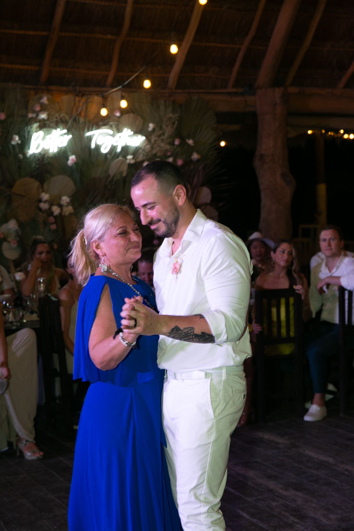 Groom in white suit shares a tender mother-son dance at tropical wedding reception.