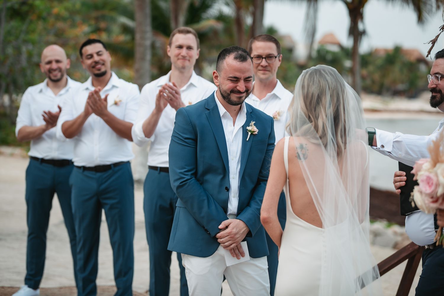 Emotional groom tears up as bride approaches during beach wedding ceremony while groomsmen applaud in background.