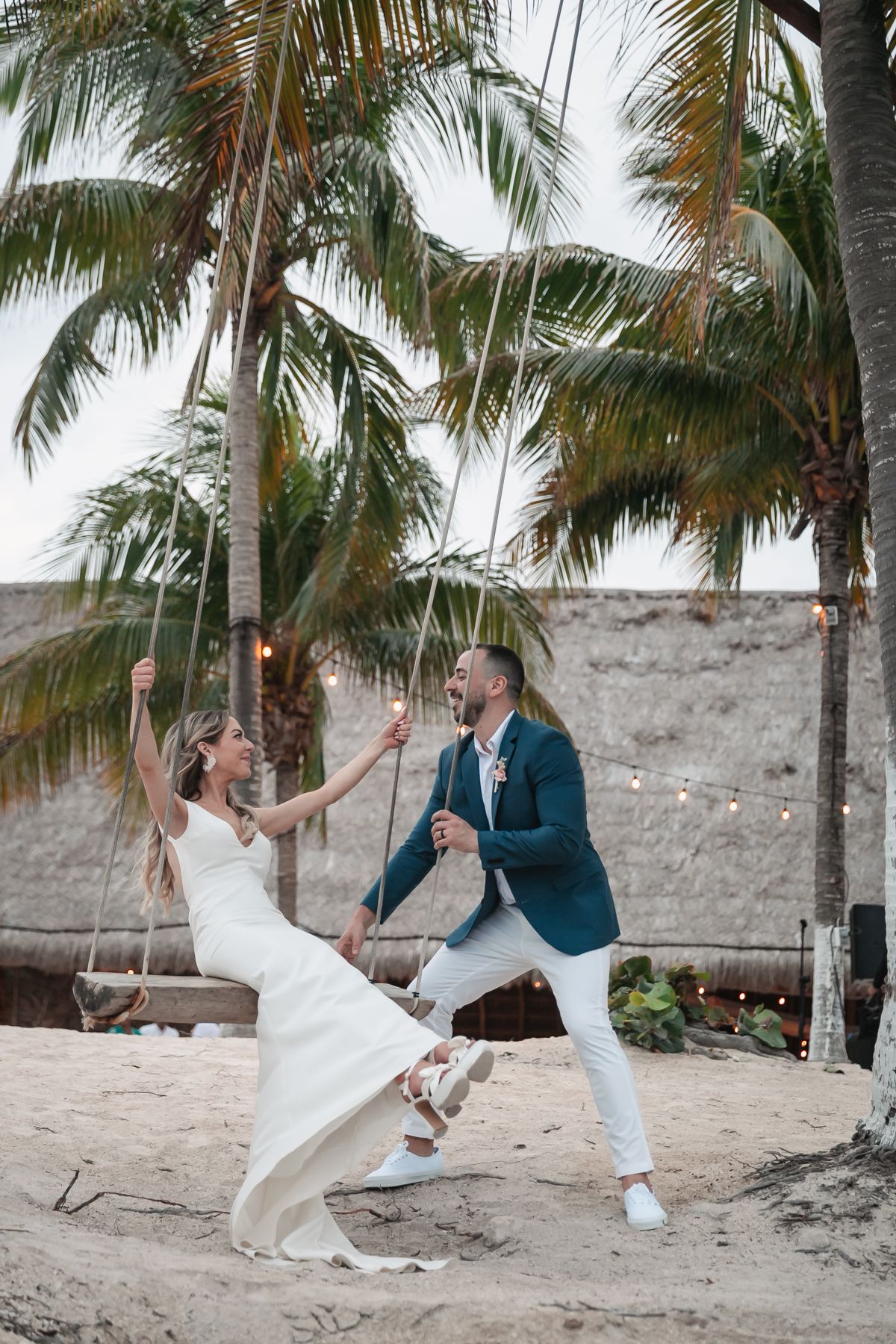 Joyful bride and groom laughing on rope swing surrounded by palm trees at tropical beach wedding.