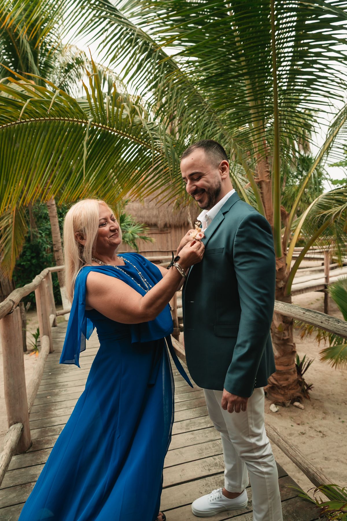 Woman in blue dress adjusts boutonniere on smiling groom in green suit on a tropical wooden bridge with palm trees.