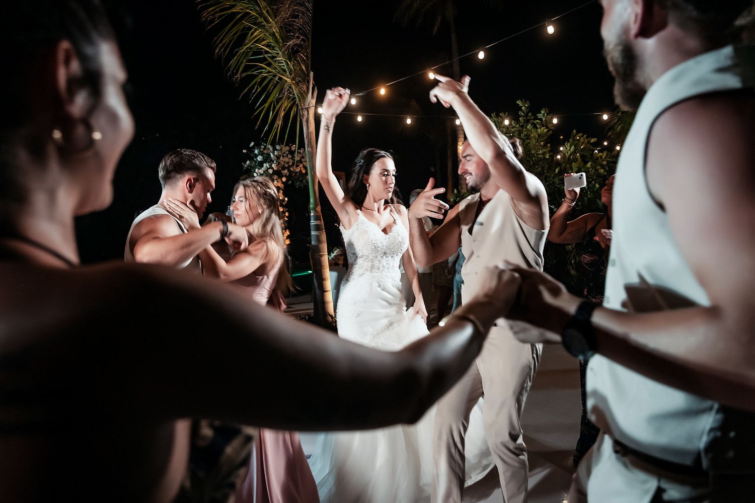 Bride and groom dancing joyfully with wedding guests at an outdoor night reception under string lights.