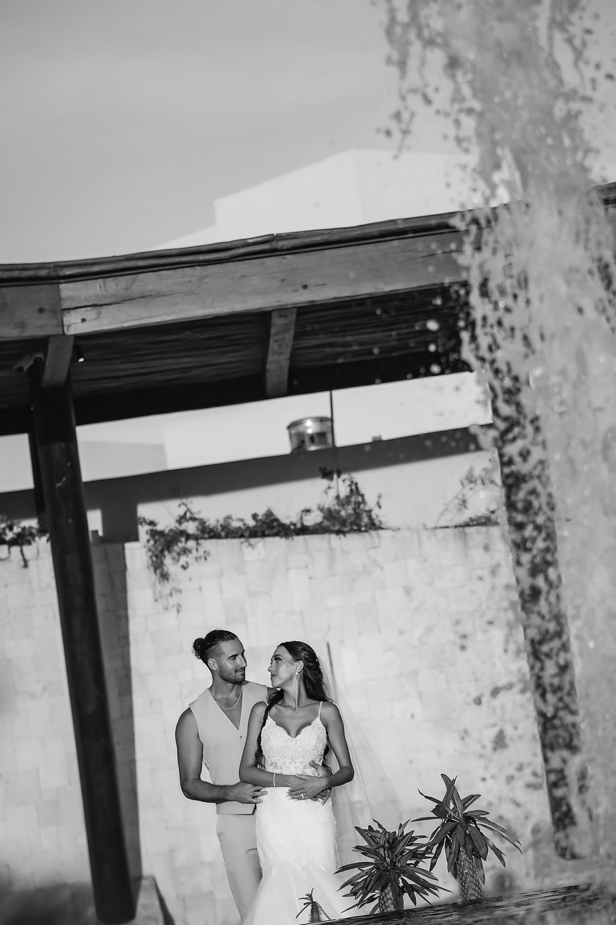 Black and white photo of a couple embracing near a fountain and tropical plants at an outdoor wedding venue.