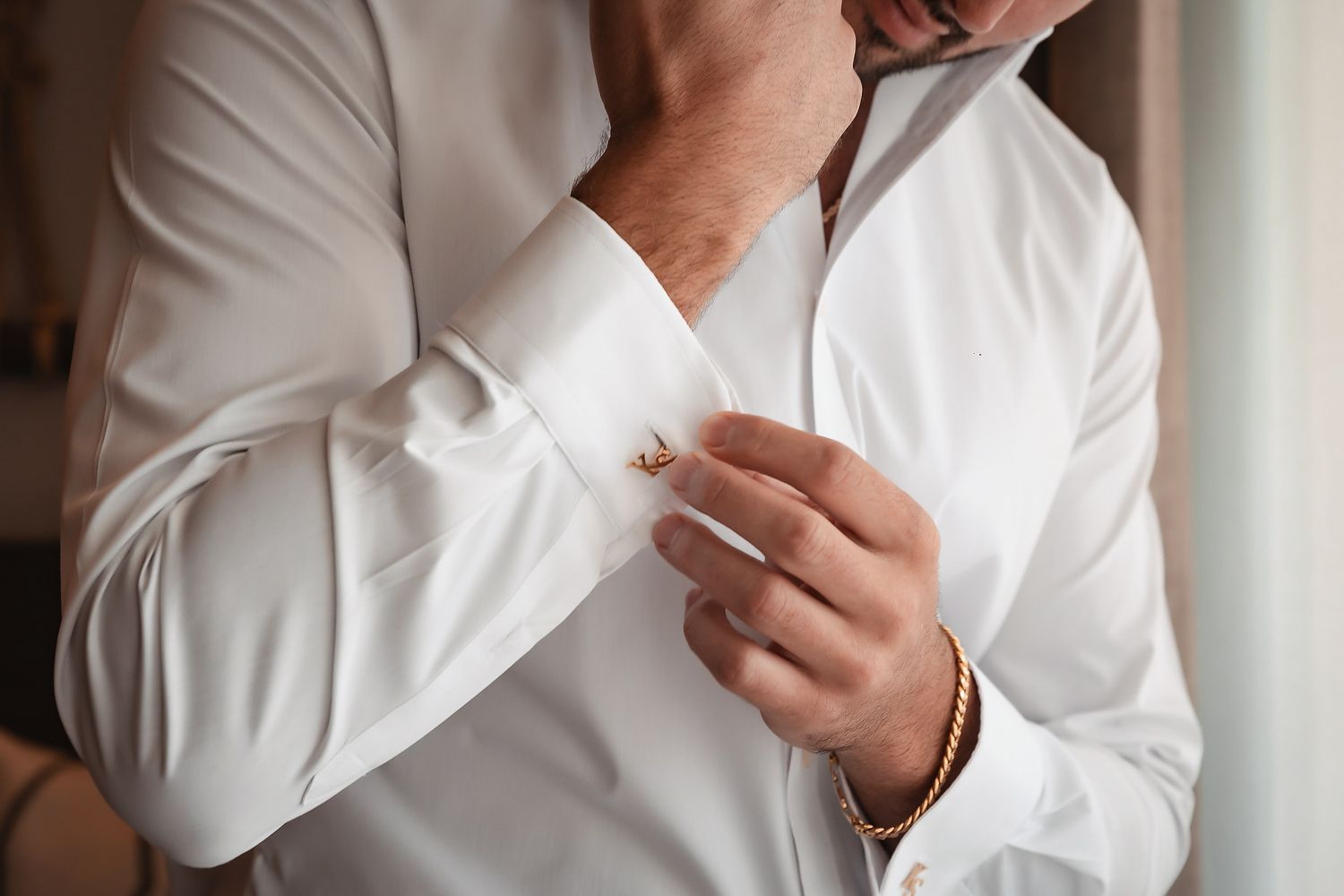 Man in white dress shirt fastening gold cufflinks on his sleeve, wearing a gold bracelet.