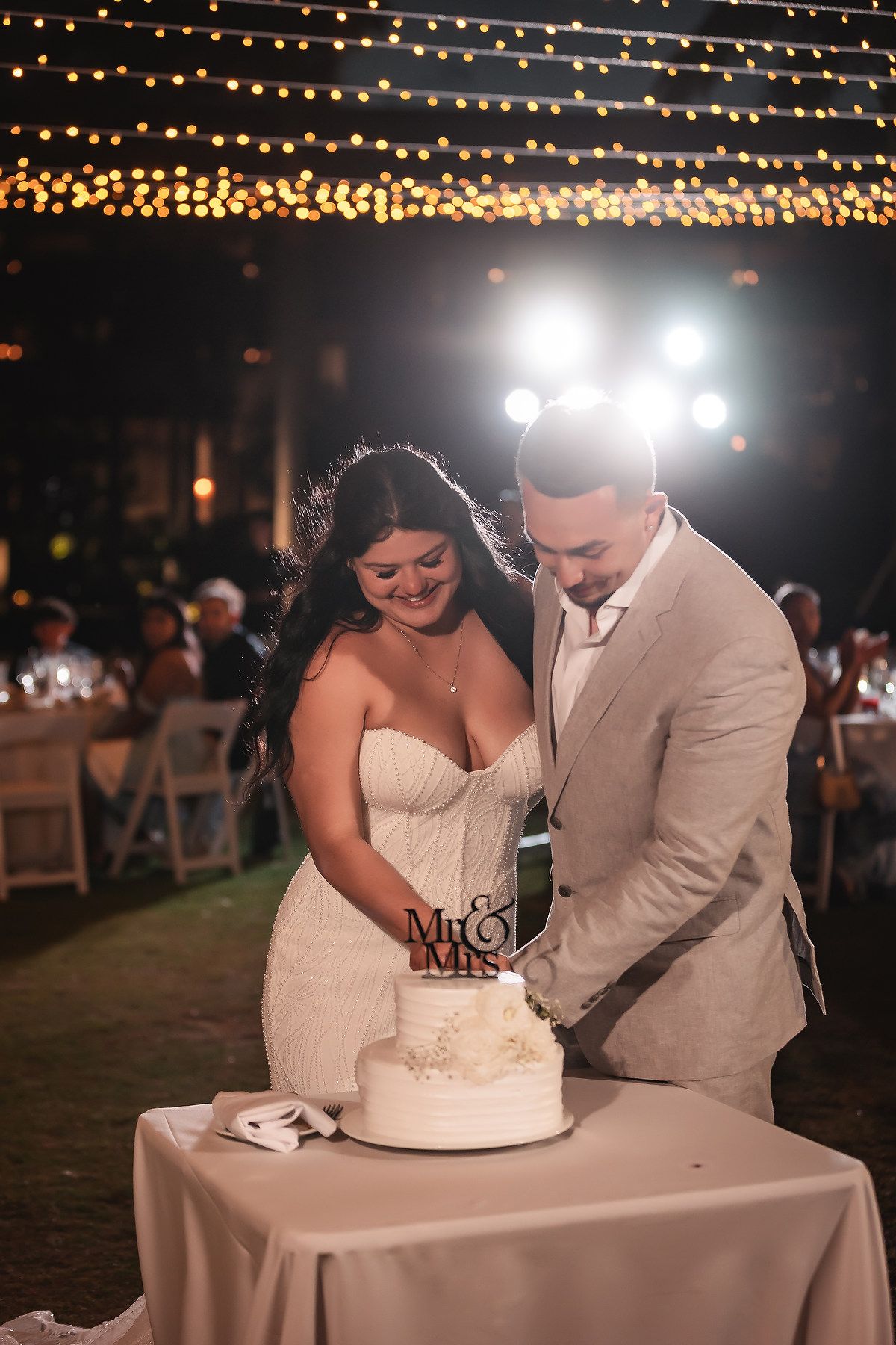 Bride and groom cutting their white wedding cake topped with Mr. & Mrs. sign at evening reception.