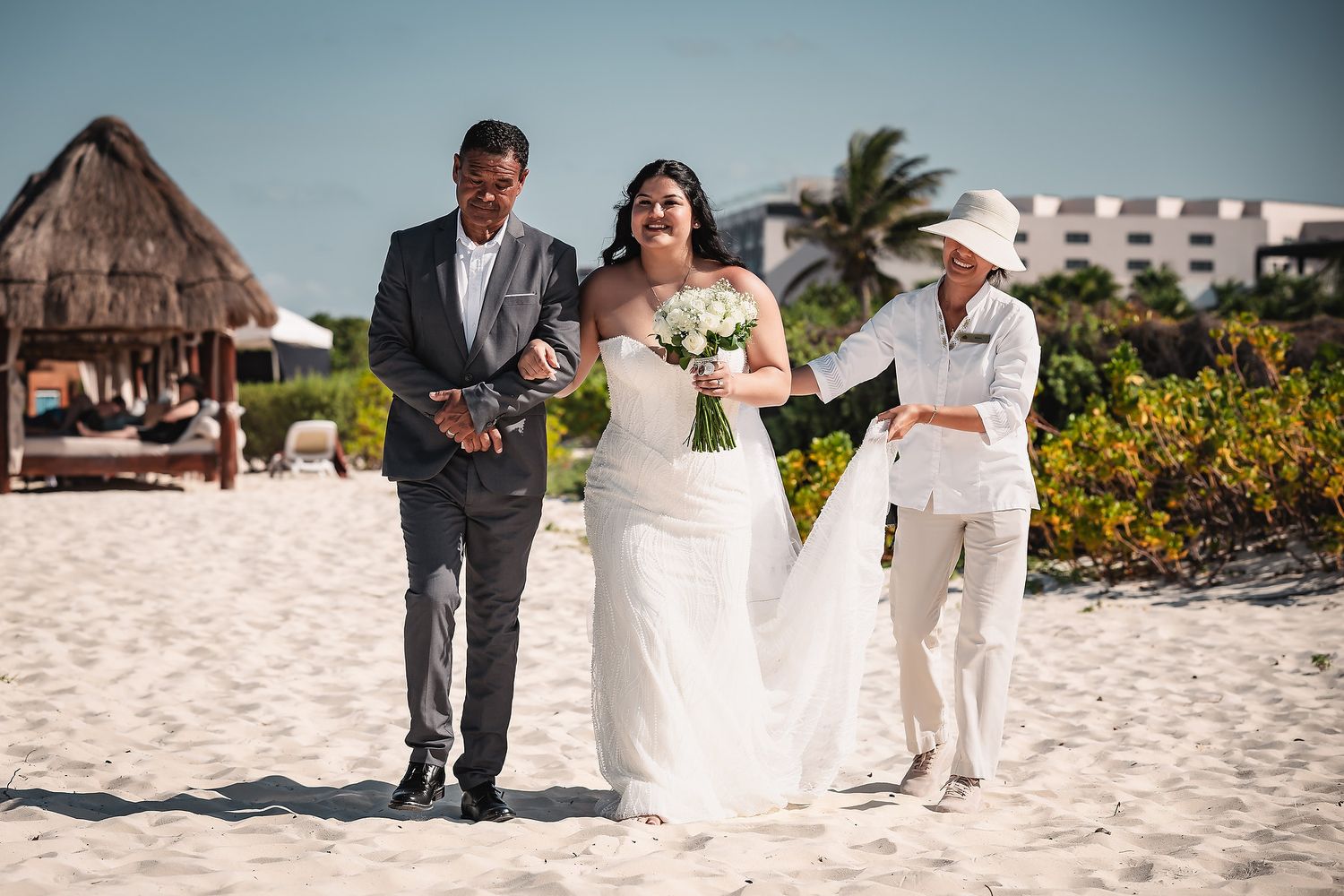 Bride in white gown walks on sandy beach with groom and officiant during outdoor wedding ceremony at tropical resort.
