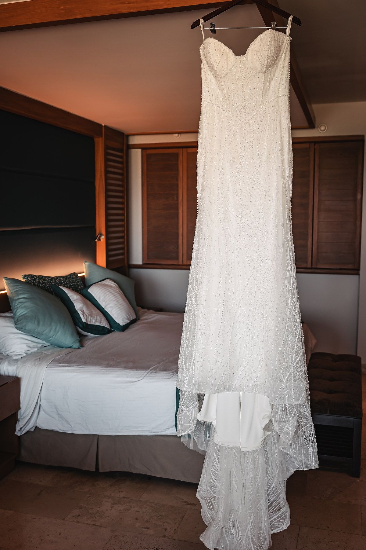White strapless lace wedding gown hanging in a warmly lit hotel bedroom with teal accent pillows.