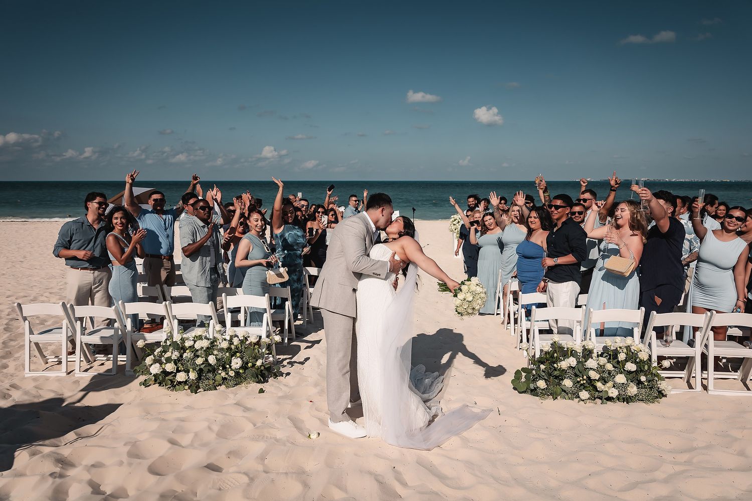 Couple shares first kiss at romantic beach wedding ceremony with guests cheering on white sandy shore under blue sky.