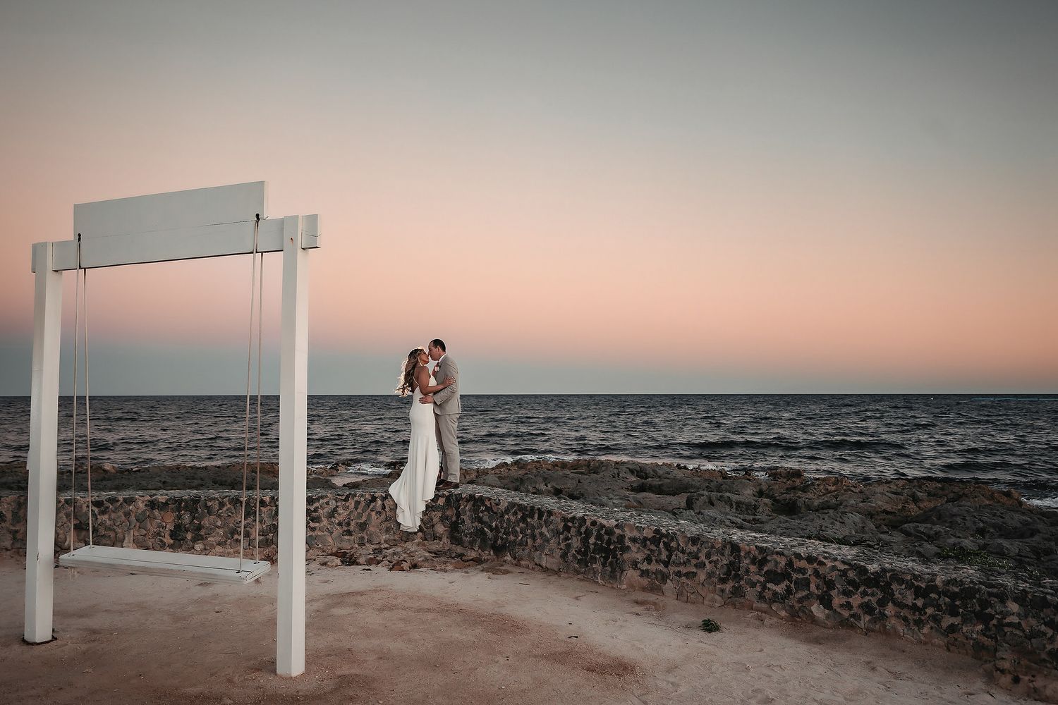 Couple embracing under a white swing arch on a rocky seaside cliff during a colorful sunset.