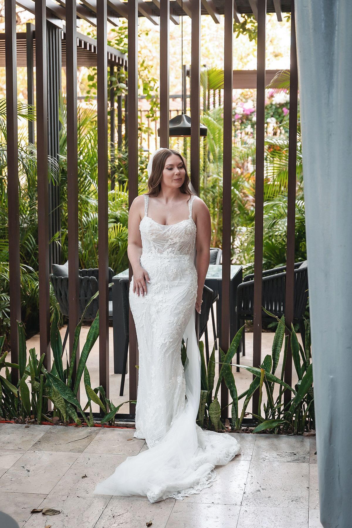 Bride in elegant white lace fitted wedding gown with train, posing outdoors among tropical greenery.
