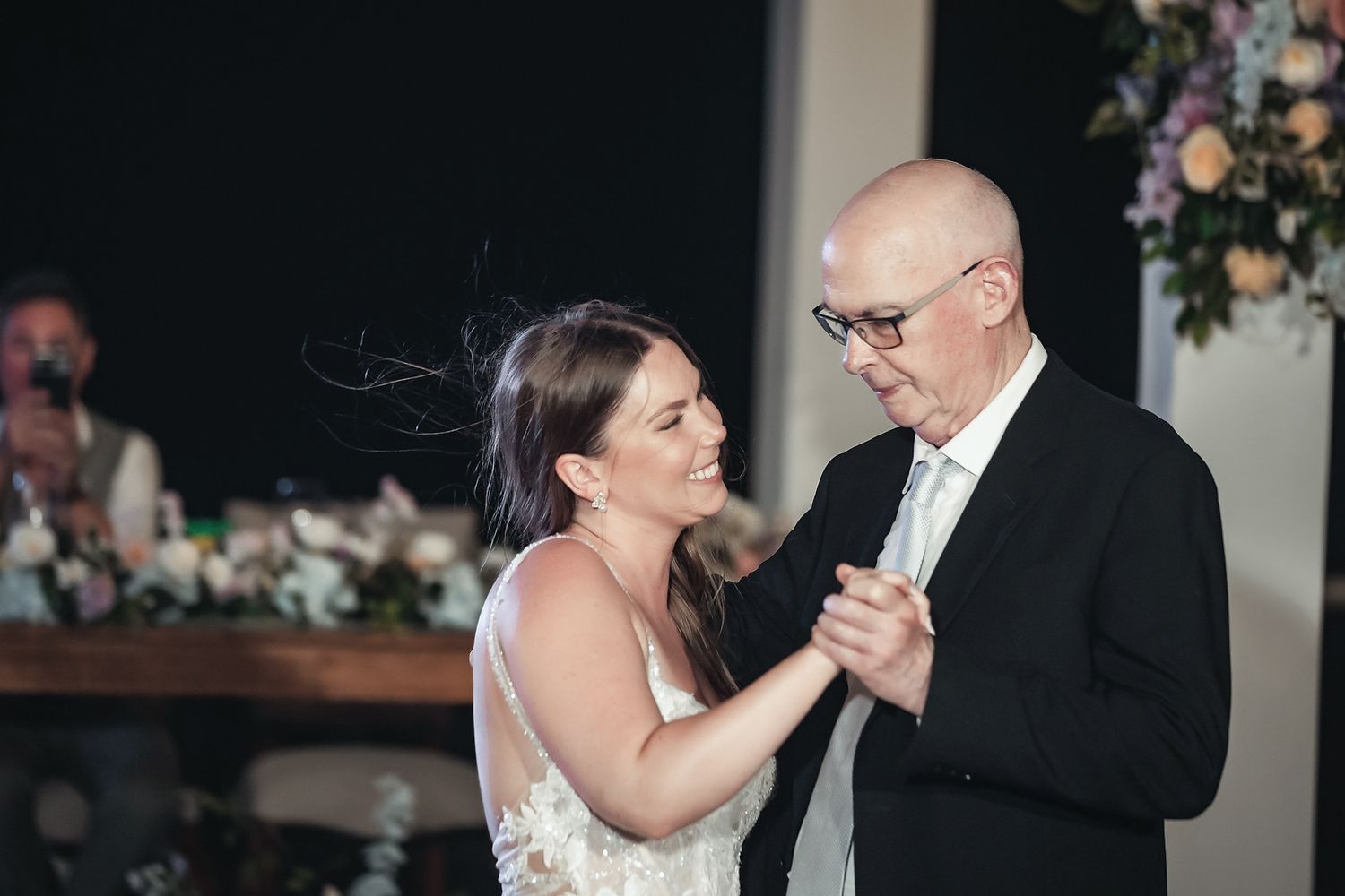 Bride and father share an emotional first dance at wedding reception with floral arrangements in background.