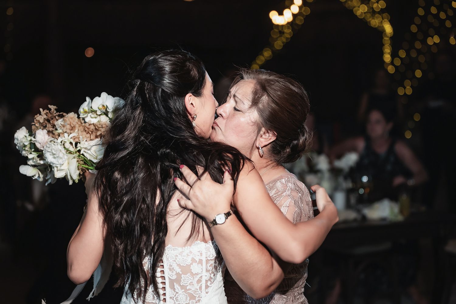 Two women share a heartfelt kiss at a wedding reception, one holding a bridal bouquet under warm string lights.