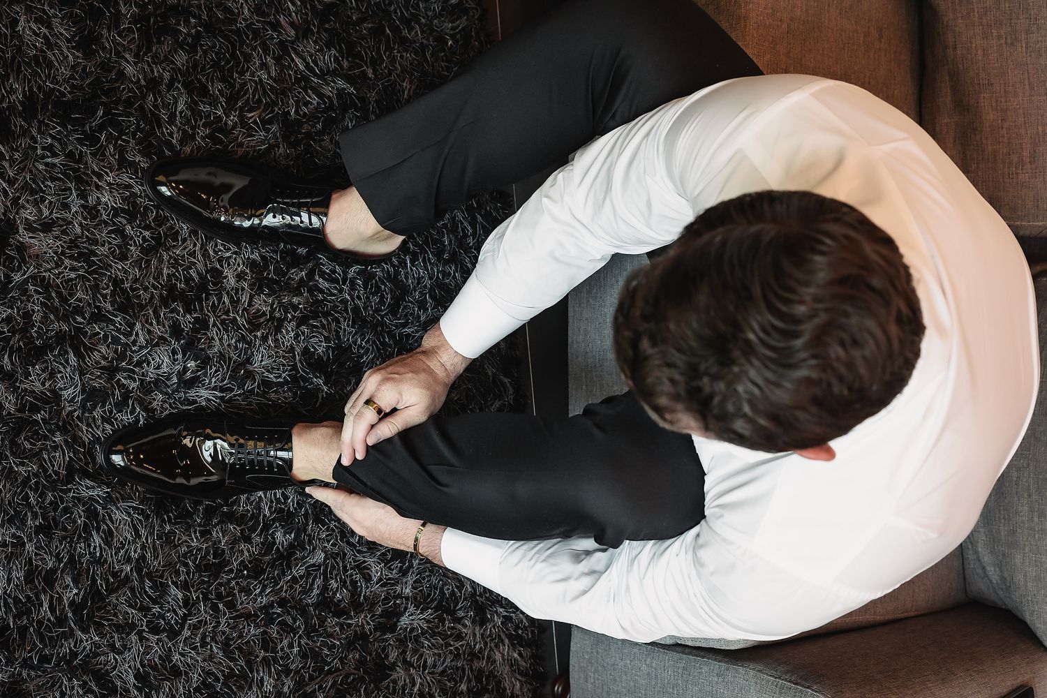 Man in white shirt and black pants tying his dress shoes while seated on a sofa, viewed from above.