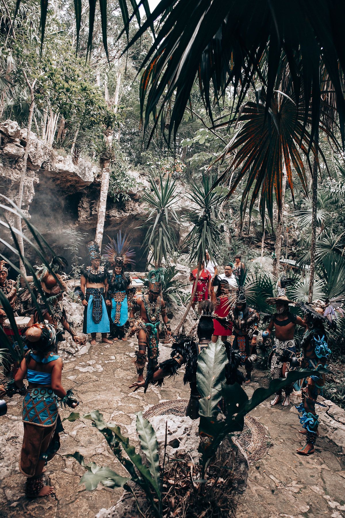 Group of performers in traditional Mayan ceremonial costumes gather in a lush jungle clearing with smoke.
