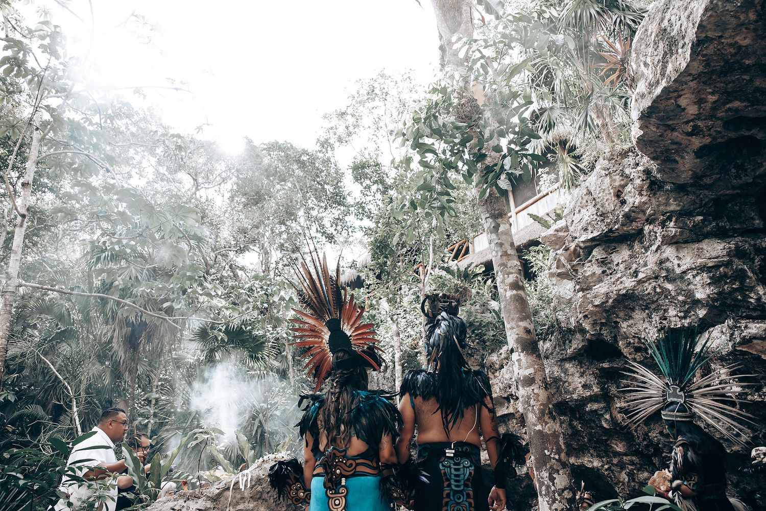 Group of Mayan ceremonial dancers in traditional feathered costumes gather near rocky cave surrounded by jungle trees.