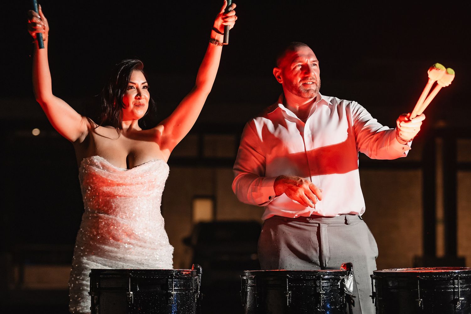Bride and groom joyfully playing large drums together at their wedding reception under warm red lighting.