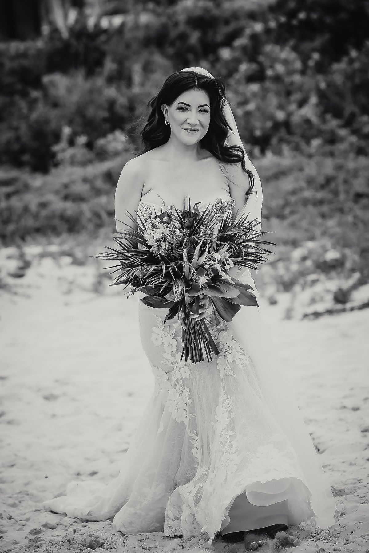 Black and white photo of a bride in a lace gown holding a tropical bouquet on a sandy beach.