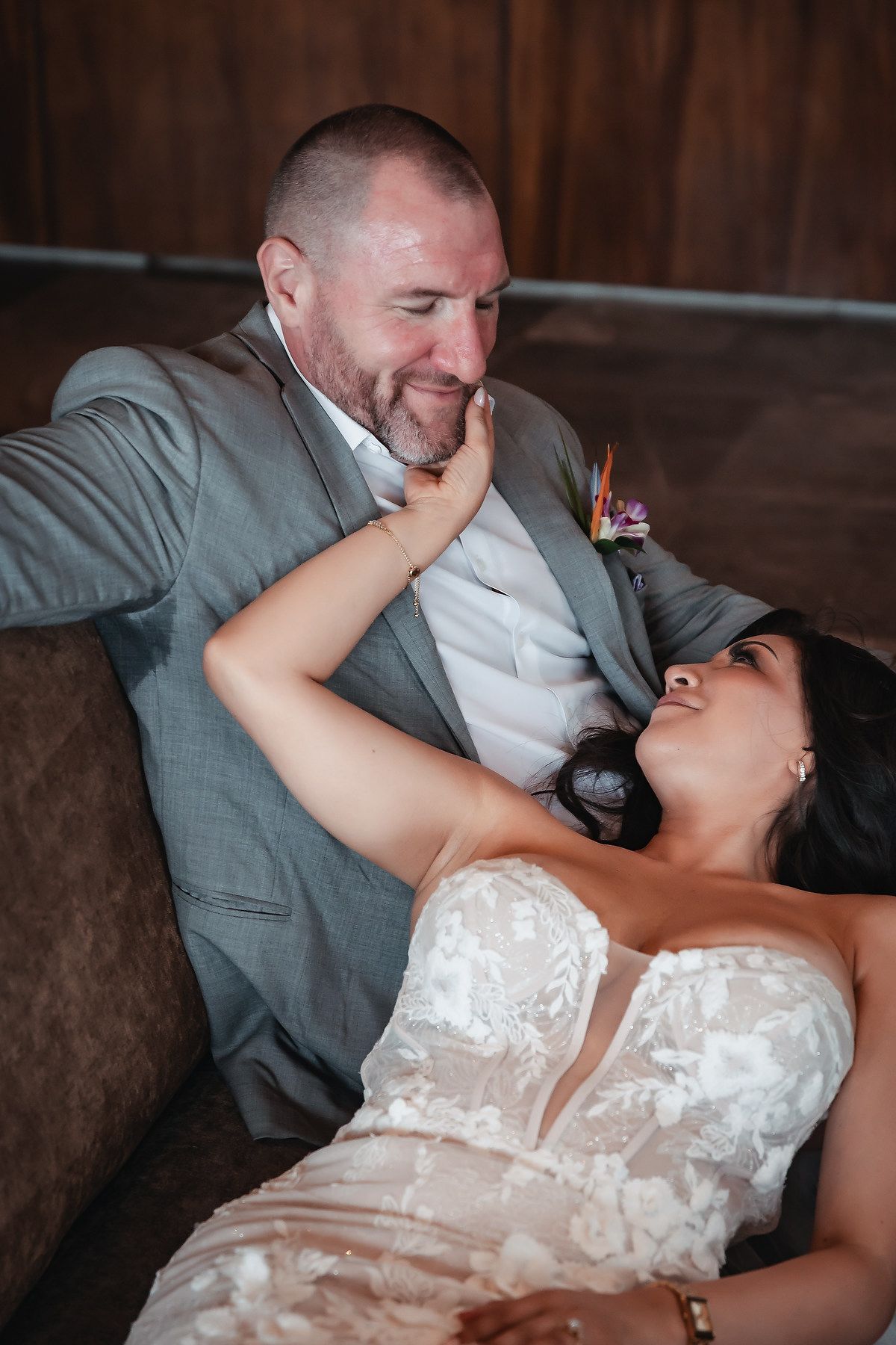 Bride gazes lovingly at groom while reclining on a sofa, gently touching his face in an intimate wedding portrait.