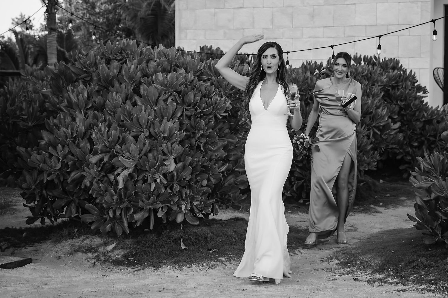 Bride in white halter gown walks outdoors with bridesmaid in gray dress amid lush greenery at beach wedding.