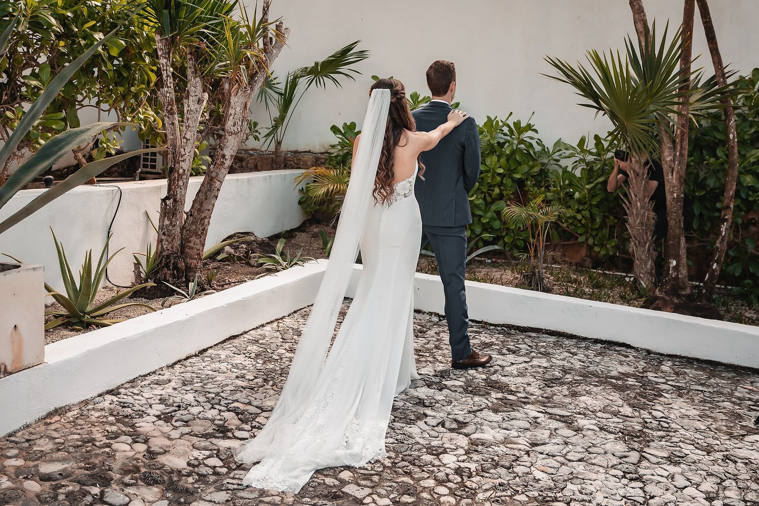 A bride in a white gown and veil stands with a groom in a blue suit in a tropical garden setting.