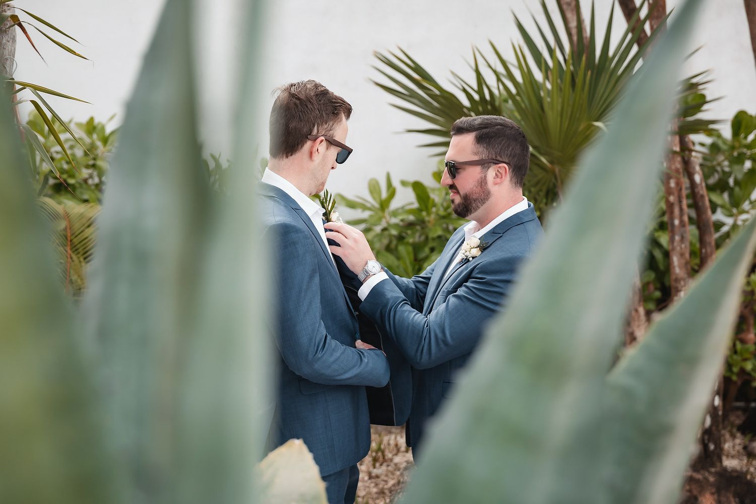 Two grooms in matching blue suits adjust ties among tropical plants before their wedding ceremony.