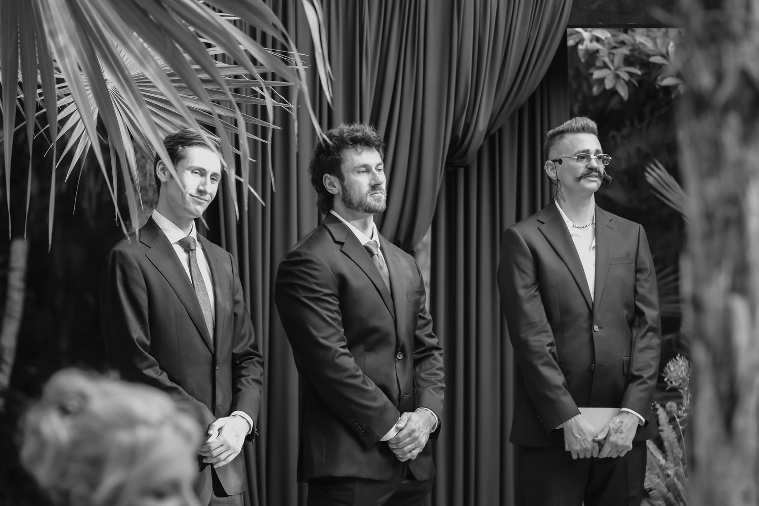 Three men in dark suits stand at wedding altar with draped curtains in black and white photo.