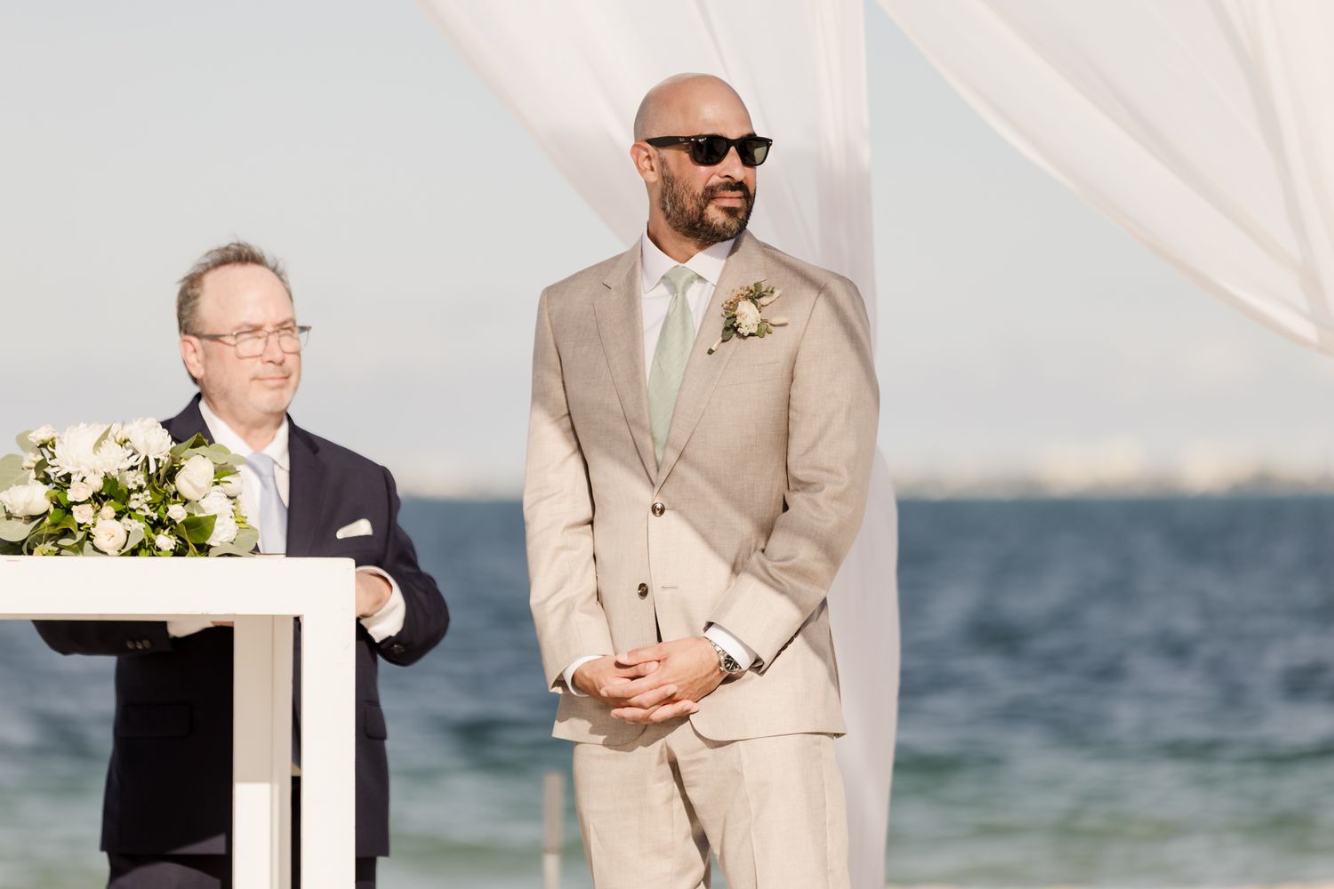 Groom in beige suit and sunglasses stands at beach wedding ceremony altar with officiant at podium.