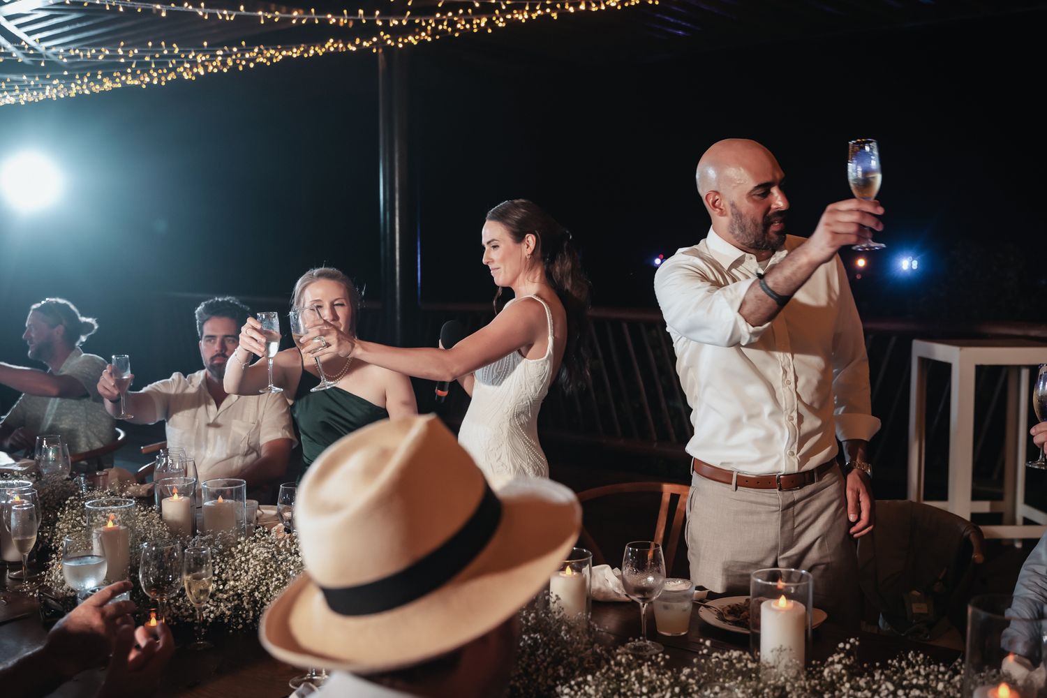 Bride and groom raising glasses in a toast with guests at an outdoor evening wedding reception with warm fairy lights.